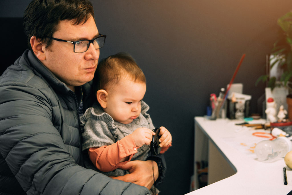 Man father with baby in carrier working at office or home with computer at the desk, parent in office